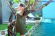 A woman in a green dress leaning against a tree by a pool.