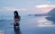 A naked woman kneeling in the water on a beach.